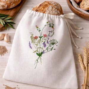 Può includere: Un sacco per il pane in lino bianco con chiusura a coulisse, ricamato con un colorato motivo floreale. La borsa contiene una pagnotta di pane croccante. Il ricamo presenta fiori e foglie rosa, viola e verdi. Un accessorio da cucina rustico e affascinante.
