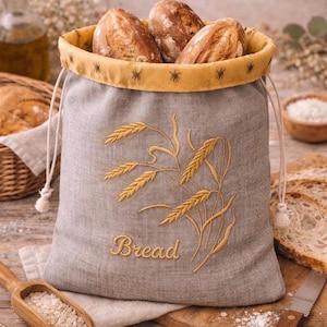 May include: A grey linen drawstring bread bag featuring golden yellow wheat stalk embroidery and the word Bread stitched in cursive. The bag has a mustard yellow interior lining with small dark star patterns and contains three crusty bread rolls.