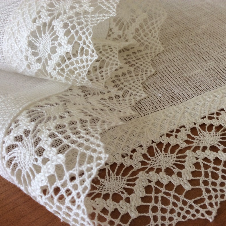 May include: White linen tablecloth with a delicate white lace trim. The lace is intricate and has a floral pattern.