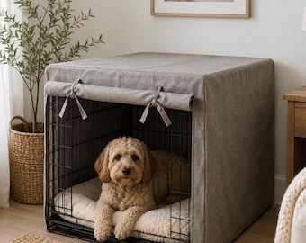 Puede incluir: Una jaula rectangular de alambre negro para perros cubierta con una funda de tela gris sólida, con un panel frontal enrollable asegurado por dos lazos de tela. Dentro, un perro de pelo rizado color café claro descansa sobre una cama mullida.