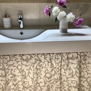 May include: A white bathroom sink with a chrome faucet and a decorative floral arrangement. The arrangement includes pink and white peonies in a silver vase. A patterned fabric skirt hangs below the sink.