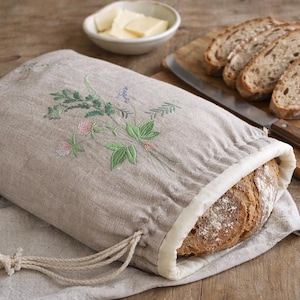 May include: A beige linen bread bag with an embroidered floral design, holding a loaf of bread. Sliced bread, a knife, and a small bowl of butter are on a wooden surface.