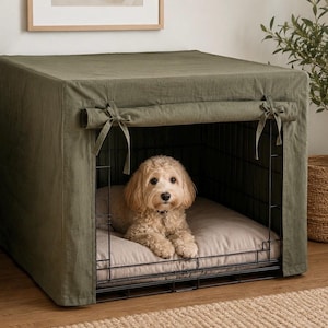 Puede incluir: Una jaula de alambre negro para perros cubierta con una funda de tela verde oliva con solapas frontales atadas. Dentro de la jaula, un cojín rectangular de color beige claro sirve de base para un perro pequeño de pelo rizado color café claro.