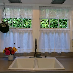 May include: A white kitchen sink with two basins, a chrome faucet, and a bouquet of flowers in a white vase. The sink is set against a backdrop of two windows with white lace curtains and green foliage visible outside.