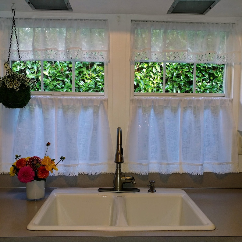May include: A white kitchen sink with two basins, a chrome faucet, and a bouquet of flowers in a white vase. The sink is set against a backdrop of two windows with white lace curtains and green foliage visible outside.