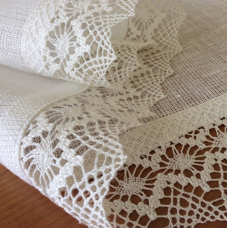 May include: White linen tablecloth with delicate white lace trim. The lace is intricate and has a floral pattern.