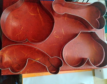 May include: A collection of four metal cookie cutters in various shapes, including a heart, a bone, and two other abstract forms. The cutters are silver-colored and rest on a textured, reddish-brown surface.