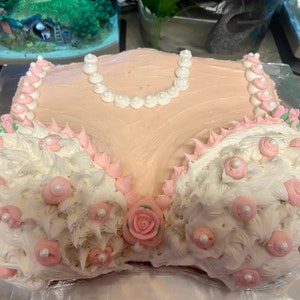 May include: A pink and white cake decorated with white frosting, pink flowers, and pearl-like decorations. The cake is shaped like a bra.