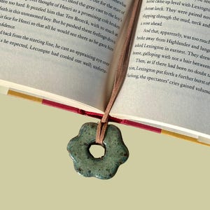 May include: A book with an open page featuring printed text and a light brown leather cord bookmark. Attached to the cord is a small, five-petaled ceramic flower charm in a mottled sage green and blue glaze with a central circular cutout.