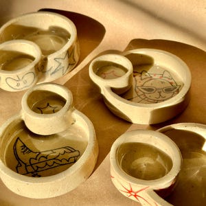 May include: A collection of handmade ceramic dishes with attached small circular containers. Each dish features unique black line art designs, including a cat wearing sunglasses, a boot, and abstract shapes. The dishes are beige with a glossy finish.