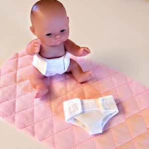 May include: A small doll with a neutral expression sits on a pink quilted mat. The doll wears a white diaper. Another white diaper is next to the doll on the mat. The doll is a toy.