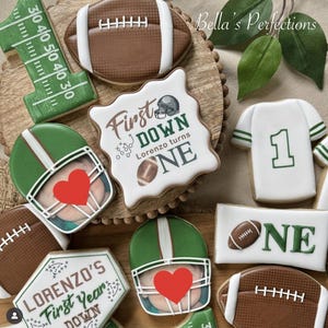 May include: A collection of decorated sugar cookies with a football theme. The cookies include footballs, helmets with hearts, a jersey with the number one, and cookies with text that reads "First Down" and "Lorenzo turns ONE".