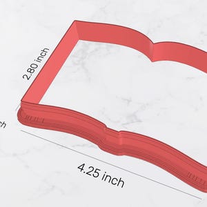 May include: Red plastic cookie cutter shaped like an open book. The cutter is 4.25 inches wide and 2.80 inches tall. The area of the cutter is 10.91 square inches.