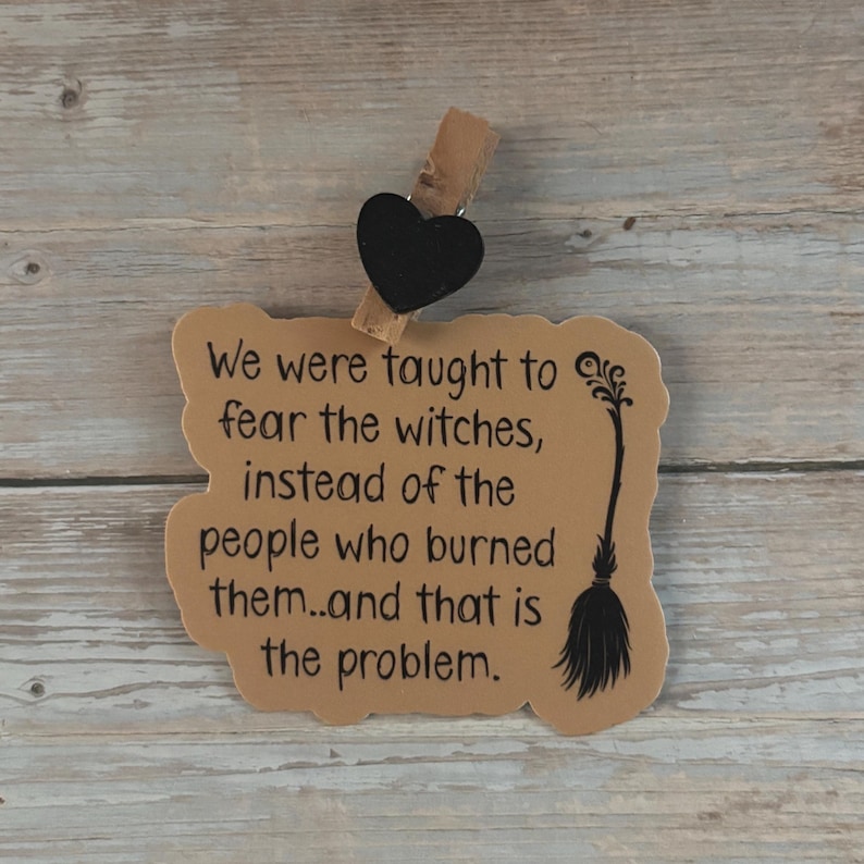 May include: A decorative sign with a message about witches and those who burned them. The sign is tan with black text and a black heart-shaped clip. A black broom graphic is on the right side of the sign.