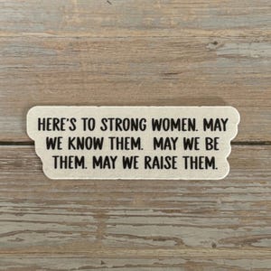 May include: A rectangular sticker with a scalloped edge, featuring the text "HERE'S TO STRONG WOMEN. MAY WE KNOW THEM. MAY WE BE THEM. MAY WE RAISE THEM." in black font. The sticker is off-white.