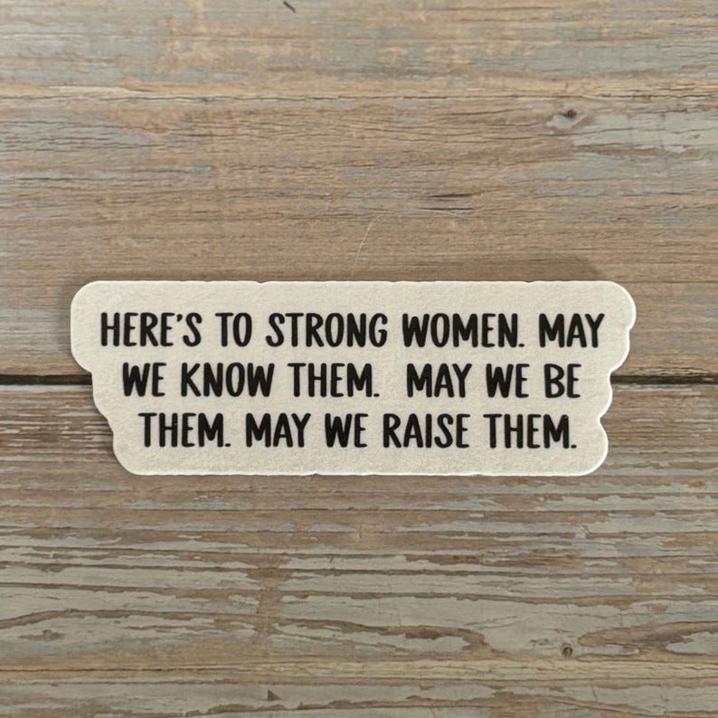 May include: A rectangular sticker with a scalloped edge, featuring the text "HERE'S TO STRONG WOMEN. MAY WE KNOW THEM. MAY WE BE THEM. MAY WE RAISE THEM." in black font. The sticker is off-white.
