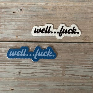 May include: Two stickers with the phrase "well...fuck." One sticker has a white background with black text, and the other has a blue background with blue text. The stickers are on a wooden surface.
