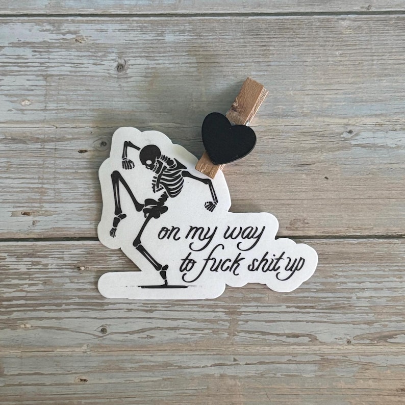 K&ouml;nnte beinhalten: Ein wei&szlig;er Aufkleber mit einer schwarzen Skelettgrafik in einer Tanzpose. Der Aufkleber tr&auml;gt den Schriftzug "on my way to fuck shit up" in Schreibschrift. Eine schwarze herzf&ouml;rmige Klammer ist befestigt.