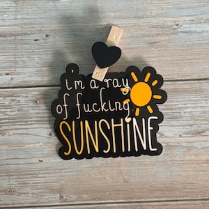 May include: A black and yellow sign with the text "I'm a ray of fucking sunshine" and a sun graphic. The sign is attached to a wooden clothespin with a black heart. The background is a light blue wooden surface.