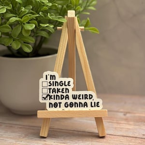 May include: A sticker on a small wooden easel with the text "I'm SINGLE, TAKEN, KINDA WEIRD, NOT GONNA LIE" in black font. The sticker has check boxes next to each option. A small potted plant is in the background.