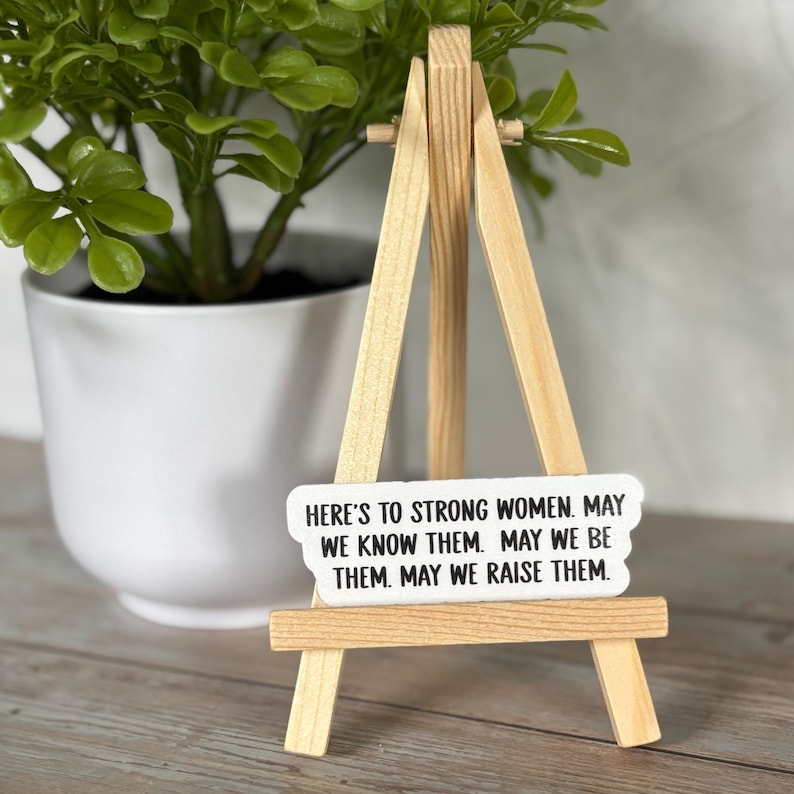 May include: A small wooden easel holds a white rectangular sign with black text. The sign reads, "HERE'S TO STRONG WOMEN. MAY WE KNOW THEM. MAY WE BE THEM. MAY WE RAISE THEM." A small potted plant is in the background.