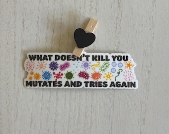 May include: A white sticker with the text "WHAT DOESN'T KILL YOU MUTATES AND TRIES AGAIN" and colourful microbe illustrations. A small black heart is attached to a wooden clothes peg, placed above the sticker.