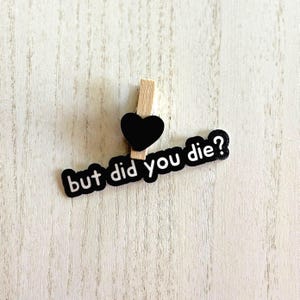 May include: A wooden clothespin with a black heart and a black and white text that reads "but did you die?" The clothespin is on a light wood-grain surface.