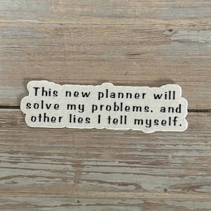 May include: A white sticker with black text that reads: "This new planner will solve my problems, and other lies I tell myself." The sticker is on a wooden surface.