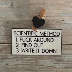 May include: A white rectangular sign with black text that reads "SCIENTIFIC METHOD 1. FUCK AROUND 2. FIND OUT 3. WRITE IT DOWN." A wooden clothespin with a black heart is attached to the top.