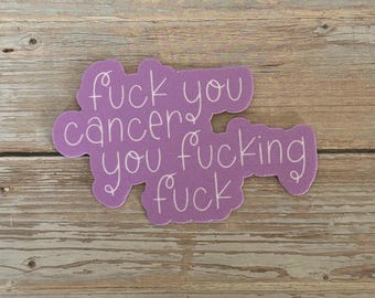 May include: A lavender sticker with the words "fuck you cancer you fucking fuck" in a handwritten style. The sticker is on a wooden surface.
