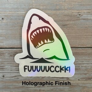 May include: Holographic sticker featuring a shark with an open mouth and the word "FUUUUUCCKK!" in black text. The sticker has a holographic finish, with colors that shift in the light. The shark is depicted in black and white.