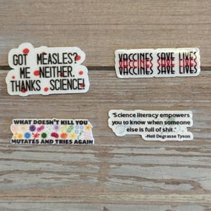 May include: Four white stickers with black text and red accents. One sticker reads "GOT MEASLES? ME NEITHER. THANKS SCIENCE!". Another says "VACCINES SAVE LIVES". A third reads "WHAT DOESN'T KILL YOU MUTATES AND TRIES AGAIN". The last says "Science literacy empowers you to know when someone else is full of shit."