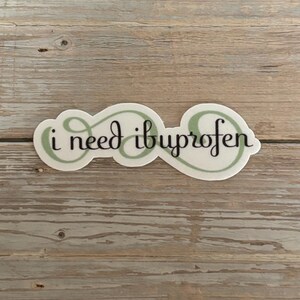 May include: A white sticker with the phrase "i need ibuprofen" in a black, cursive font. The text is set against a light green, swirly background. The sticker is rectangular with rounded edges.