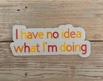 May include: A white sticker with the phrase "I have no idea what I'm doing" in orange, yellow, and red lettering. The sticker is on a light brown wooden surface.