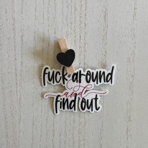 May include: A white sticker with the phrase "fuck around and find out" in black and burgundy script. A small black heart is attached to a wooden clothespin, which is clipped to the sticker. The sticker is on a light wood-grain surface.