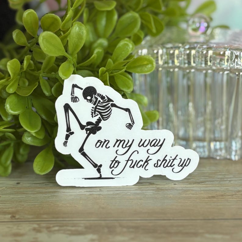 K&ouml;nnte beinhalten: Ein wei&szlig;er Aufkleber mit einem schwarzen Skelett in einer dynamischen Pose. Der Text "on my way to fuck shit up" ist in kursiver Schrift darunter geschrieben. Der Aufkleber liegt auf einer Holzoberfl&auml;che.