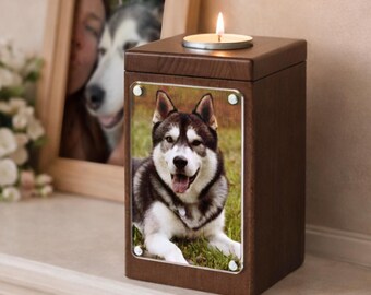 May include: A wooden pet urn with a photo of a Husky dog on the front. The urn has a small metal plate and a candle on top. A framed photo of a dog and flowers are in the background.