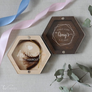 May include: Two hexagonal wooden keepsake boxes. One light-colored box contains a lock of hair with the text "Andrew's FIRST HAIRCUT 4.17.2023". The other dark-stained box lid features the text "FIRST CURL Amy's 4.17.2023" inside a geometric frame.