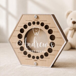 May include: A hexagonal wooden keepsake box for baby teeth featuring a clear acrylic front panel. The surface is engraved with a tooth icon, the name Andrew's, and the text FIRST TEETH, along with circular slots for teeth labeled UPPER and LOWER.