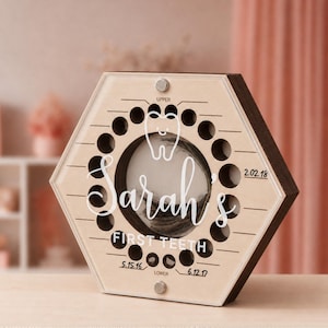 Könnte beinhalten: Eine sechseckige Zahn-Aufbewahrungsbox aus Holz mit einer transparenten Acrylfront. Sie verfügt über ein zentrales kreisförmiges Fach, ein weißes Zahnsymbol, den Namen Sarah's, den Text FIRST TEETH sowie Beschriftungen für UPPER und LOWER mit den Daten 2.02.18, 5.15.16 und 6.12.17.