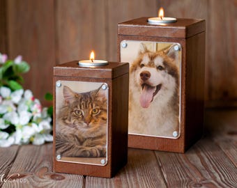 May include: Two wooden pet memorial boxes with lit candles on top. One box features a cat photo, the other a dog. The boxes are brown with clear acrylic photo covers, and are set on a wooden surface.