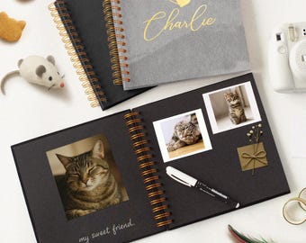 May include: An open black photo album with cat photos, a pen, and the text "my sweet friend." Two closed albums are visible, one black and one gray with the name "Charlie" in gold script. Cat-shaped treats and a toy mouse are also present.