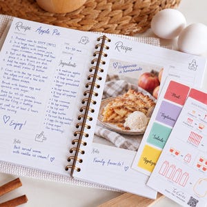 May include: A spiral-bound recipe book open to an Apple Pie recipe page with printed instructions, ingredients, and a photograph of a slice of pie with ice cream. A separate card with kitchen measurement conversions and category tabs is placed on top.