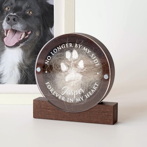 May include: A round, clear glass keepsake with a brown wooden base. The glass contains a paw print and fur from a pet. The inscription reads "No longer by my side, Forever in my heart, Jasper".