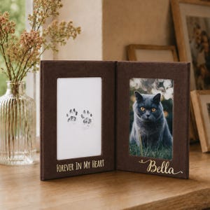 May include: A dark brown, hinged double photo frame with a velvet-like texture. The left side displays two black paw prints and the text "FOREVER IN MY HEART" in gold. The right side holds a photograph of a grey cat with the gold name "Bella".
