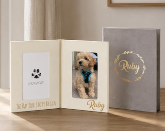 May include: A cream-colored photo frame with a picture of a puppy and a paw print. The frame has the text "THE DAY OUR STORY BEGAN" and the puppy's name, "Ruby". A gray photo album with "Ruby" in gold script is next to the frame.