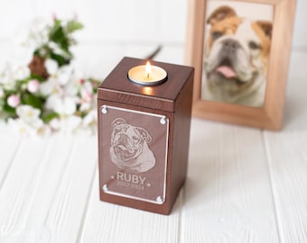 May include: A brown wooden pet memorial urn with a lit candle on top. The front of the urn features an engraved image of a bulldog and the name "Ruby" with the years 2007-2024. A framed photo of a bulldog is in the background.