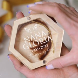 May include: A wooden hexagon-shaped keepsake box with a clear lid, containing a lock of brown hair and a gold chain. The box is engraved with the name "NANA GLORIA", dates, and the phrase "forever in my heart".