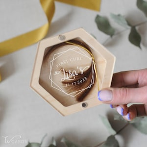May include: A hexagonal light wood keepsake box with a clear acrylic lid featuring white printed text that reads "FIRST CURL", "Lisa's", and "11.17.2023" inside a geometric frame. A lock of brown hair is visible inside the wooden container.