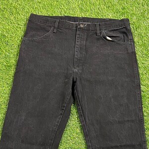 May include: Black denim jeans with a classic five-pocket design. The jeans are made of a sturdy material and feature a button and zip closure. The jeans are laid flat on a green surface.